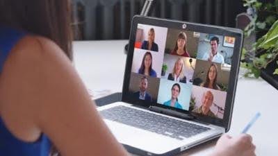 Closeup of a laptop on a table while a female watches participants of the video conference on screen