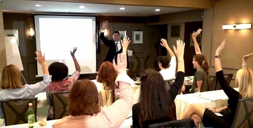 Dr. Kousouli stands at the front of the class in a black suit and dress pants teaching a corporate company wellness healing class in Los Angeles, CA New York, NY Miami, FL