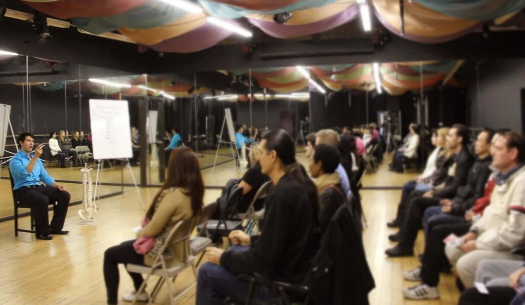 Dr. Kousouli sitting at the front of the class in a blue shirt and dress pants teaching a hypnosis class in Los Angeles, CA New York, NY Miami, FL